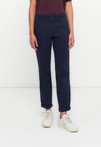 Ženska v temnopiščenih hlačah, ki so zvite, belih supergah in temno vijoličasti majici s kratkimi rokavi, stoji ob belem ozadju.