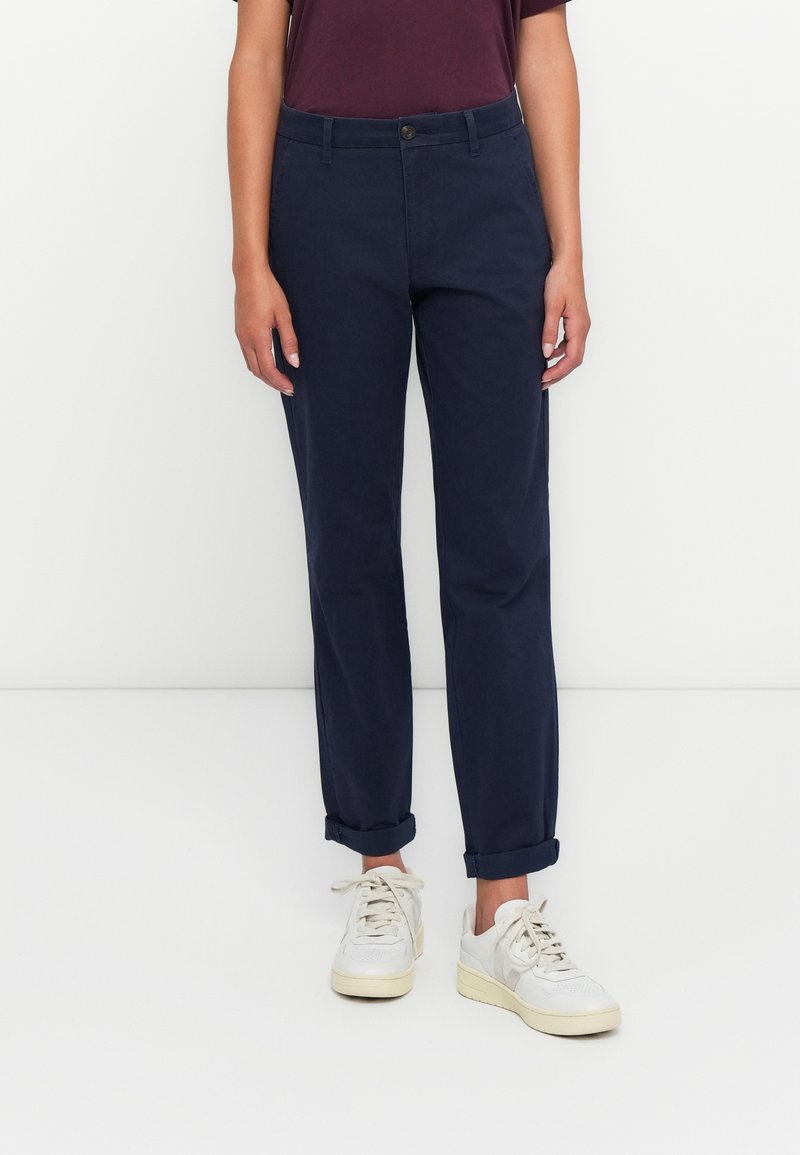 Ženska v temnopiščenih hlačah, ki so zvite, belih supergah in temno vijoličasti majici s kratkimi rokavi, stoji ob belem ozadju.