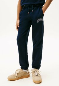 Navyblauwe sweatpants met elastische boorden, een trekkoord in de taille en "TOMMY" geborduurd in het wit op het linkerbeen. Draag ze met tan sneakers.