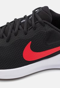 Svart sportskor med en strukturerad mesh-övre, röd Nike-logotyp, svarta snörning och en vit gummisula.