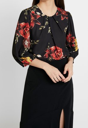 Frau trägt eine schwarze Bluse mit großem roten und gelbem Blumenmuster und 3/4-Puffärmeln, kombiniert mit einem schwarzen Rock mit einem vorderen Schlitz.