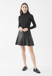 Maglietta nera a maniche lunghe con colletto alto, abbinata a una gonna in pelle nera con pieghe. Il modello indossa mocassini neri. Sfondo semplice e pulito.