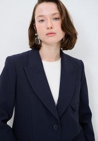 Mörkblå skräddarsydd kavaj med slagkrage, enkel knäppning och bröstficka. Tillverkad av ett slätt tyg med strukturerade axlar.