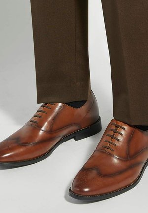 Chaussures habillées en cuir marron avec détails brogue portées avec un pantalon marron foncé et des chaussettes noires sur un fond clair.