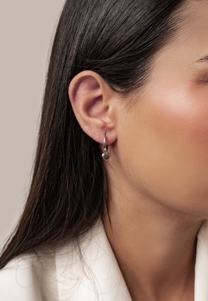 Woman's right ear with straight dark hair wearing a small silver hoop earring with a heart-shaped charm against a neutral background.