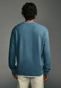 Chemise bleue texturée à manches longues avec un col rond, des poignets côtelés et un détail au dos. Associée à un pantalon décontracté de couleur claire.