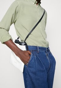 Borsa bianca in pelle con dettagli neri, texture morbida e chiusura a coulisse, indossata a tracolla sopra una camicia verde chiaro e jeans blu.