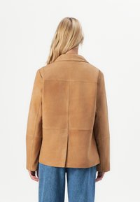 Lys hård person som bærer en tan suede-jakke med bagsyede detaljer og blå jeans, stående mod en hvid baggrund.
