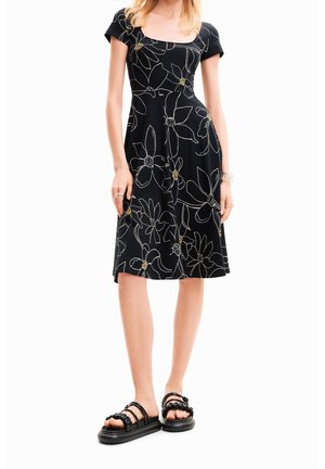 Žena v černých šatech po kolena s velkými bílými a žlutými květinovými motivy a černých sandálech s otevřenou špičkou a řetízkovými pásky.