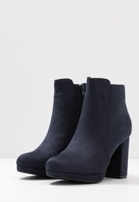 Pair of black suede ankle boots with block heels and side zippers on a white surface and background.