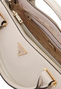 Sac à main en cuir beige avec ferrures dorées, présentant un logo triangulaire et des fermetures à glissière. L'intérieur est décoré du motif "Guess".