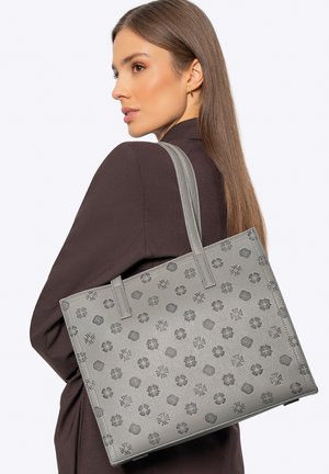 Žena s rovnými hnědými vlasy, oblečená v tmavém saku, drží na rameni šedou tašku s vzorem na bílém pozadí.