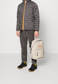 Giacca grigia imbottita con design trapuntato, maglione marrone, pantaloni in velluto a coste scuri, scarpe nere e uno zaino beige chiaro con logo rosso.