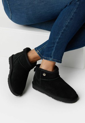 Stivali alla caviglia in suede neri con punta arrotondata, altezze corte e dettaglio a bottone. Indossati con jeans blu, che mostrano una texture liscia e suola in gomma.
