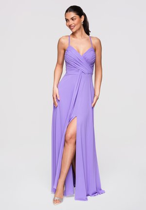 Femme portant une robe lavande à fines bretelles, corsage froncé et fente haute sur la cuisse, accessoirisée avec des boucles d’oreilles et des talons scintillants.