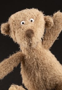 Stuffed bear toy in light brown, made of textured fabric. Features round eyes, a snout, and raised arms, with fuzzy detailing.