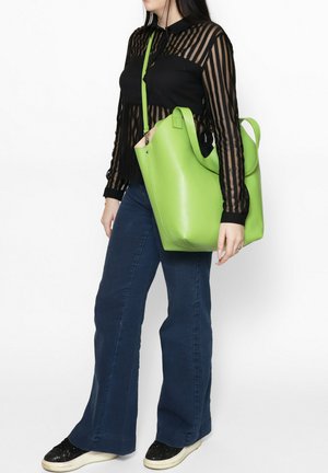 Donna che indossa una camicetta trasparente nera a strisce, jeans blu, sneakers nere, con una grande borsa in pelle verde brillante a tracolla.