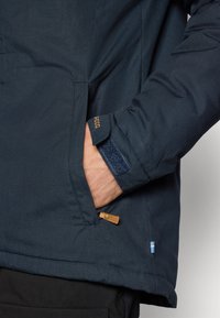 Navy blue jacket with a textured fabric, featuring a side pocket, leather accent on the zipper, and a small flag emblem at the hem.