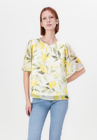 Hvit bluse med mønster av gule sitroner og grønne blader, korte puffermer, rund halslinning og lett, gjennomsiktig stoff. Brukt sammen med blå jeans.