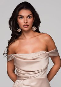 Nainen, jolla on tummat hiukset, pukeutuneena vaaleanpunaiseen satiiniseen off-shoulder-mekkoon ja suuriin kukka korvakoruihin, poseeraa vaaleanharmaata taustaa vasten.