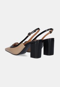 Zapatos de tacón tipo slingback en cuero beige y negro con punta afilada, que presentan un tacón grueso negro y un acento circular negro en la correa.