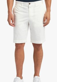 Uomo che indossa pantaloni corti bianchi fino al ginocchio e sneaker bianche, in piedi con le braccia rilassate ai lati su uno sfondo neutro.