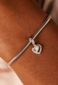 Zilveren slangenarmband met een hartvormige bedel, voorzien van een heldere steen omringd door kleine heldere accenten. Textuur van huid op de achtergrond.