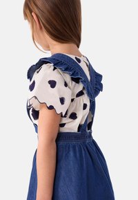 Salopette en denim bleu foncé avec des bretelles à volants sur une chemise blanche à motifs de cœurs bleu marine et aux bords festonnés.