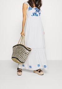 Vrouw in een witte maxi-jurk met blauwe bladpatronen, die een geweven strozak vasthoudt en zwarte plateausandalen draagt, tegen een effen achtergrond.