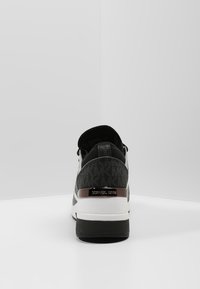 Zapatilla de Michael Kors en blanco y negro vista desde atrás, mostrando la placa del logo en el talón y la parte superior de tejido estampado.