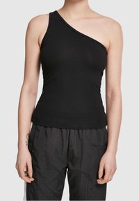 Personne portant un haut noir asymétrique sans manches et un pantalon noir ample, debout avec les bras détendus le long du corps sur un fond gris clair.