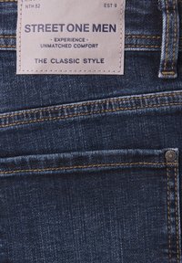 Jean en denim bleu foncé avec une étiquette beige portant l'inscription « Street One Men - Expérience de confort inégalée - Le style classique » au-dessus de la poche arrière.
