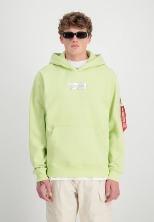 Giovane uomo che indossa occhiali da sole neri, una felpa verde chiaro con tasca frontale e etichetta rossa sulla manica, e pantaloni cargo beige su uno sfondo bianco.