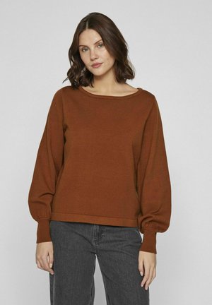 Pull en laine brun avec un col rond, des manches longues surdimensionnées et des poignets côtelés. Porté avec un pantalon gris, sur un fond uni.