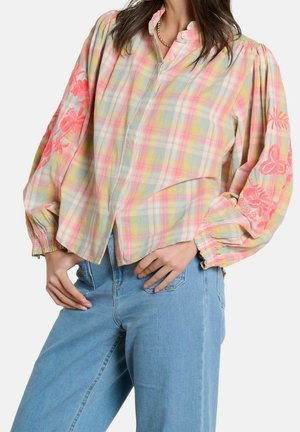 Femme portant un chemisier à carreaux pastel avec des manches bouffantes et une broderie florale rose, associé à un jean bleu clair.