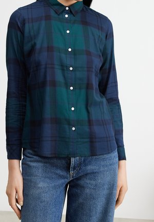 Kobieta ubrana w koszulę w zielono-granatową kratę z długimi rękawami i niebieskie dżinsy, stojąca na tle białego tła.
