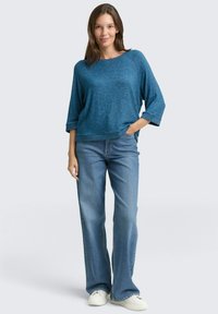 Türkise Strickjacke mit Dreiviertel-Ärmeln, kombiniert mit hellblauen Flare-Jeans und weißen Turnschuhen. Weicher Stoff mit lässiger Passform.