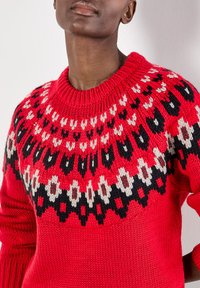 Roter Strickpullover mit rundem Ausschnitt, der einen gemusterten Einsatz in Schwarz, Weiß und Creme aufweist. Das strukturierte Garn verleiht ein lebendiges Erscheinungsbild.