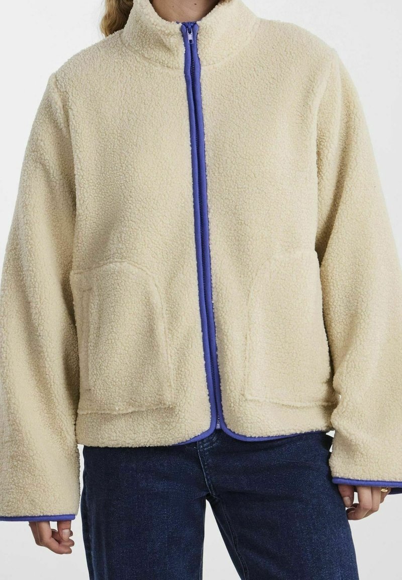 Personne portant une veste polaire couleur crème avec une fermeture éclair et des finitions violettes, associée à un jean bleu foncé, visible du cou jusqu'aux hanches.