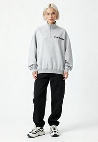 Grijze sweater met rits en de tekst "C’est la vie" in het zwart, gecombineerd met losse zwarte cargobroek en zwart-witte sneakers.