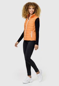 Oranges, gesteppte Weste mit hohem Kragen, weißem Reißverschluss und Seitentaschen, getragen über einem schwarzen Langarmoberteil, kombiniert mit schwarzen Jeans und weißen Sneakers.