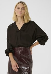 Zwarte zijden blouse met een V-hals en oversized mouwen, gecombineerd met een glanzende bordeauxrode rok met een textuurafwerking.