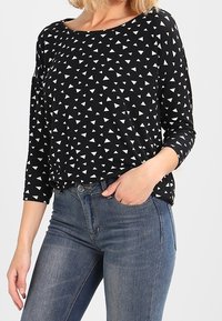 Haut noir avec un motif géométrique de triangles blancs. Le design présente un large décolleté et des manches trois-quarts. Associé à un jean ajusté.