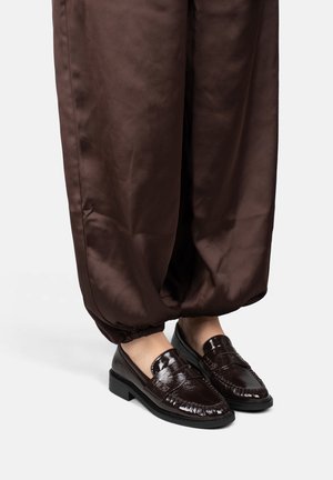 Donkere, glanzende leren loafers gedragen met losse, opgerolde bruine broek op een lichte achtergrond, waarbij voeten en enkels zichtbaar zijn.