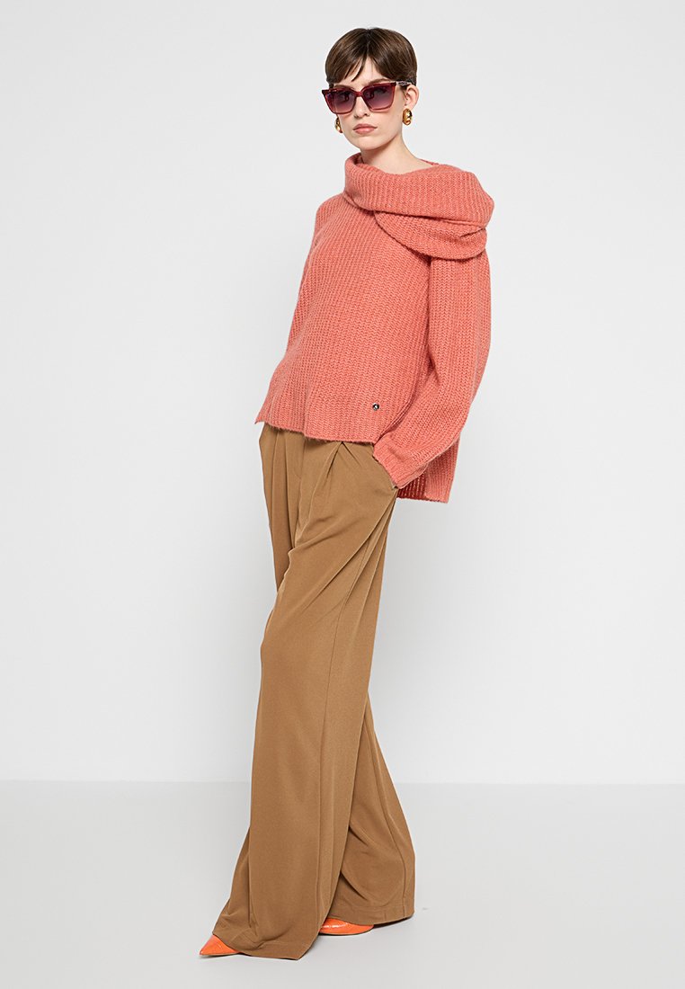 Maglione rosa lavorato a maglia con scollatura drappeggiata, abbinato a pantaloni beige a gamba larga. Il modello indossa accessori, tra cui occhiali da sole e tacchi arancioni.