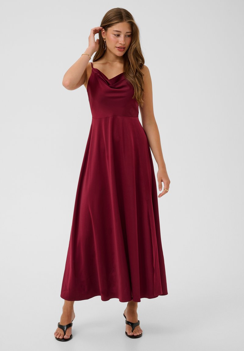 Jeune femme portant une robe en satin bordeaux sans manches avec une jupe longue jusqu'aux chevilles et des sandales noires à bout ouvert, debout devant un fond uni.