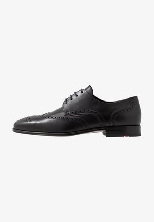 Chaussure brogue à bout golf en cuir noir pour homme avec perforations décoratives et lacets noirs, vue de côté sur fond blanc.