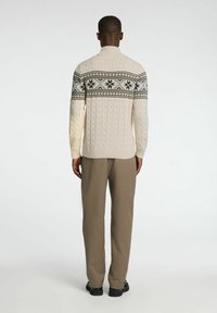 Krämfärgad kabelstickad tröja med hög krage, med ett geometriskt mönster i svart och vitt över bröstet; matchad med beige byxor.