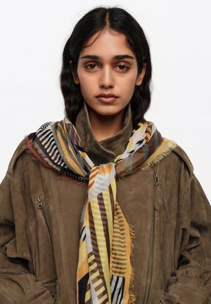 Mujer joven con cabello oscuro, vestida con una chaqueta de ante marrón y una bufanda de rayas multicolores, frente a un fondo blanco.