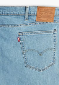 Helle blaue Denim-Jeans mit einem weißen Nähmuster und einem Leder-Brandpatch in der Taille, sowie einem roten Tab am Gesäßtasche.